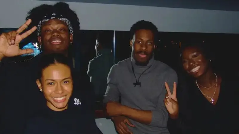 Four friends posing indoors, smiling and making peace signs, with casual attire and cheerful expressions