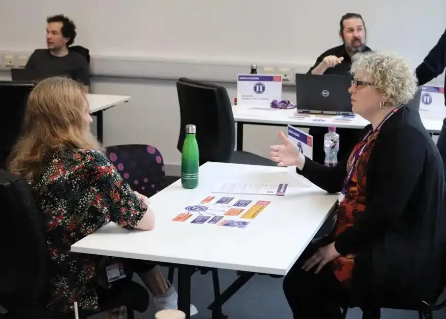 Two women seated at a table are engaged in conversation, with documents and a green water bottle in front of them. In the background, two other people work on laptops in a classroom or office setting.