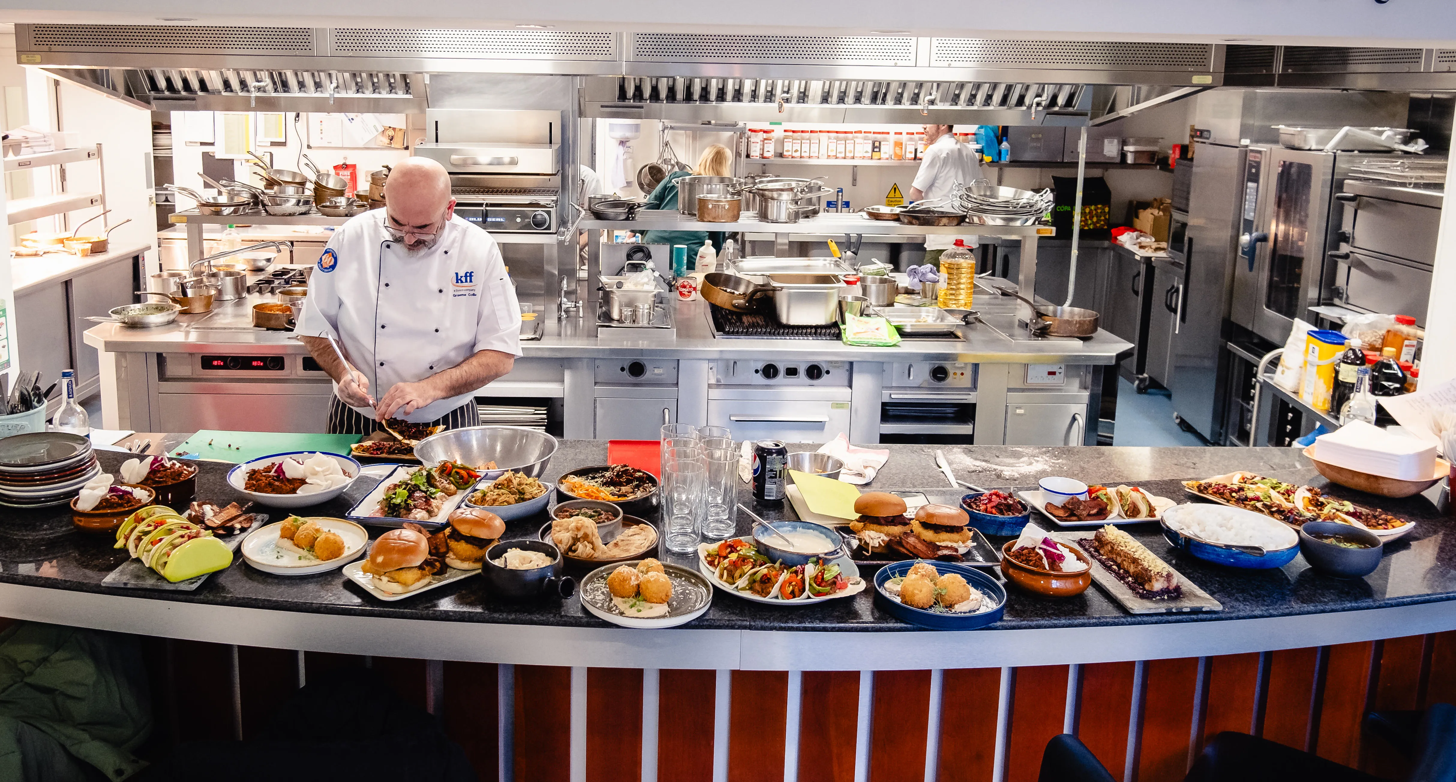 A commercial kitchen, with a table of prepared dishes including burgers