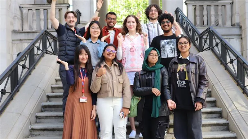 A diverse group of people smiling and posing on outdoor stairs, with a formal building and greenery in the background.