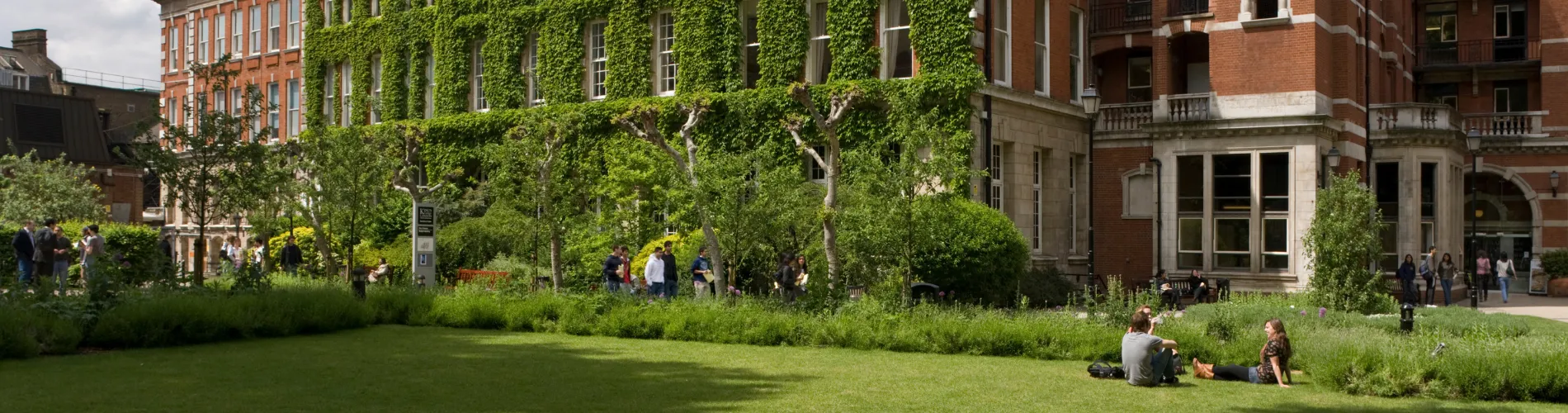 A vibrant university garden with ivy-covered buildings, students relaxing on green lawns, and tree-lined walkways under a bright, sunny sky.