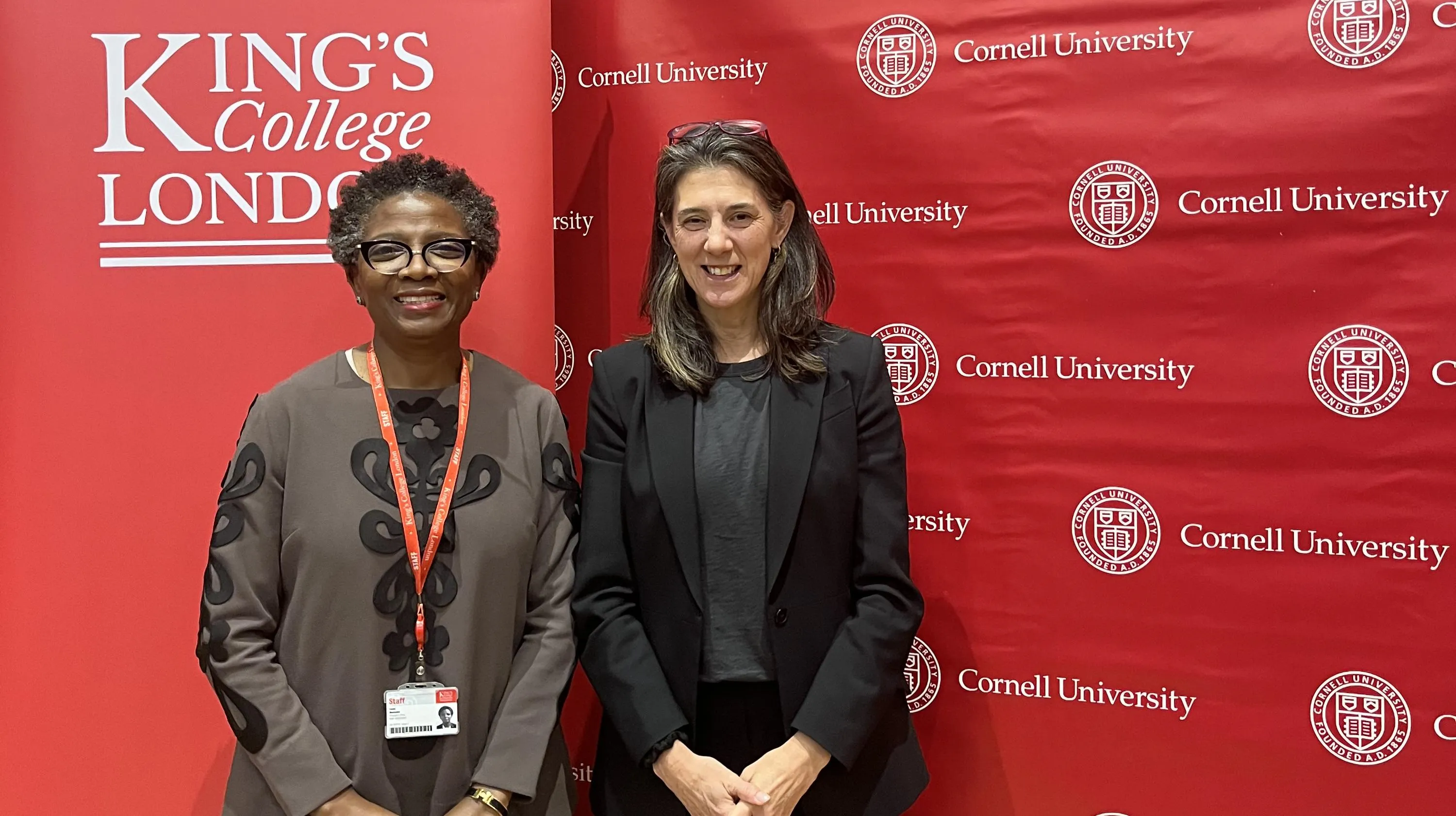 Two women, 'Funmi Olonisakin and Wendy Wolford, stand in front of a red background branded with King's College London and Cornell logos. 