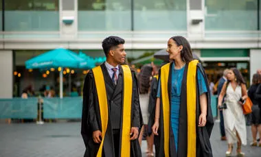 two King's graduates in black graduation gowns with yellow lining walking on the southbank talking to each other