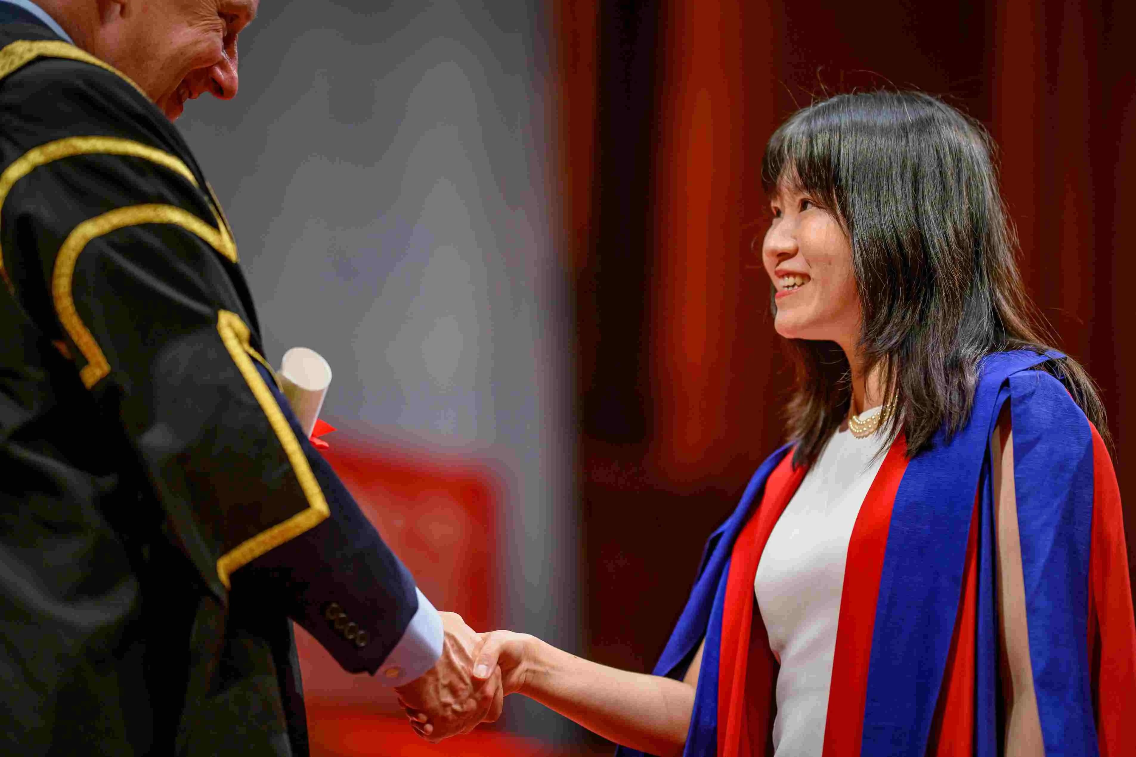 A King's graduate in a red gown with blue lining shaking the Chair's hand on stage at their graduation ceremony.