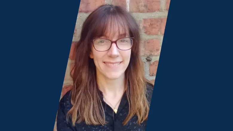 A woman with longer brown hair, a fringe and glasses smiles against a background of a red brick wall.