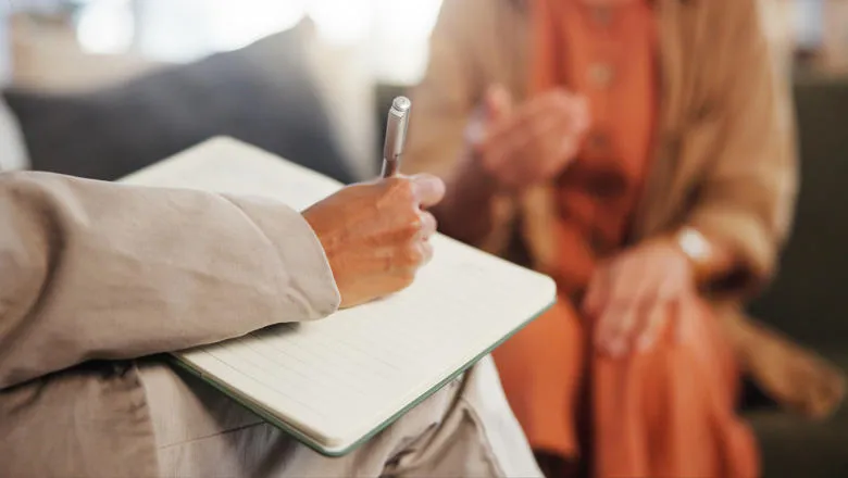 A person writing on a note pad as another person speaks in the background