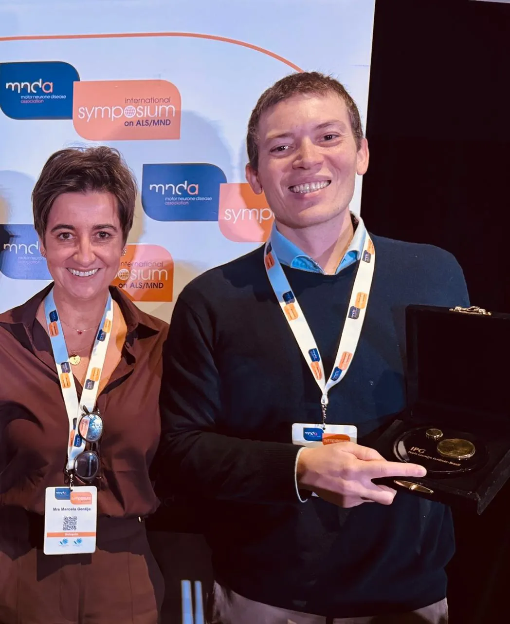 Two people stood next to each other - a women on the left and Dr Alfredo Iacoangeli on the right holding an award in an open box. the centre. They are both wearing white lanyards with MNDA symposium branded banners behind them