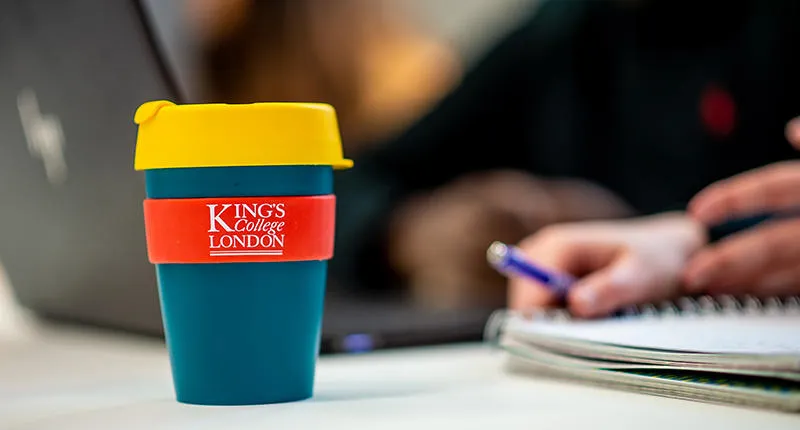 A reusable coffee cup with the King's logo, in front of a laptop and notebook