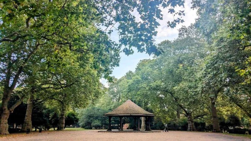 Shaded park with a central gazebo surrounded by tall, leafy trees, open pathways, and a serene atmosphere on a bright day