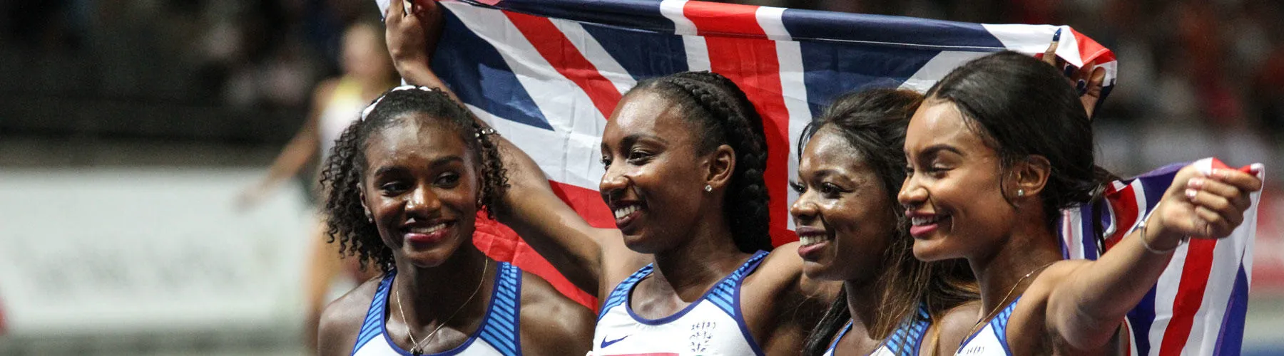 The Team GB women's relay team celebrate victory, four women hold up a flag behind them