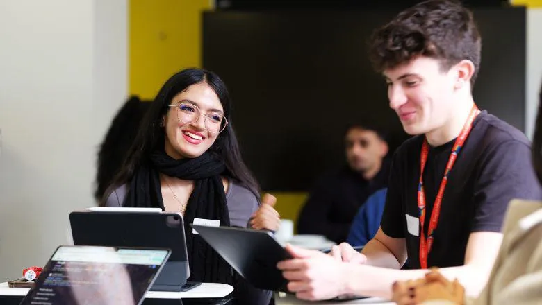 Two students are seated and smiling, working on laptops in a classroom.