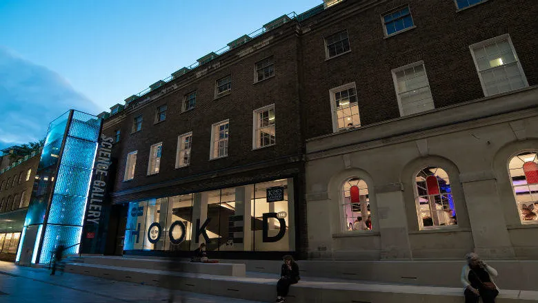 The Science Gallery London building on Great Maze Pond in the twilight, partly blue illuminated and with people sitting on the steps in front.