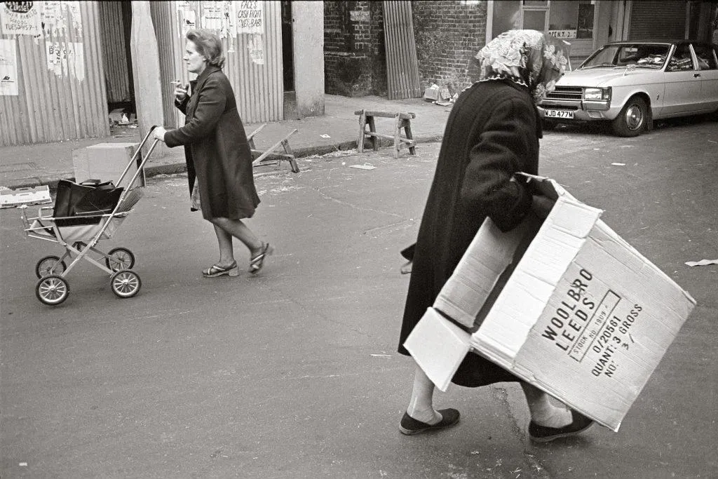 Black and white photo of a woman carrying a cardboard box and a woman pushing a pram in the background
