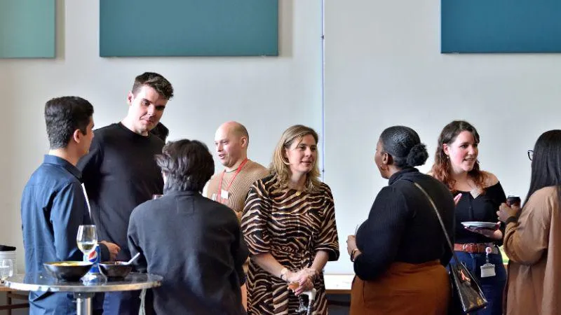 A group of people standing in a well-lit room, engaged in conversation during a social or networking event. One person in a black sweater is talking to another person in a zebra-print dress, while others, dressed in casual attire, are holding plates and d