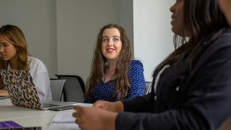 Three students sitting at a table during a discussion. A student in the center is smiling while looking slightly to the side. Two other students, one with a laptop, are focused on their work,