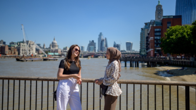 Two students talking on a bridge over the Thames with St Paul's Cathedral and Canary Wharf in the background.