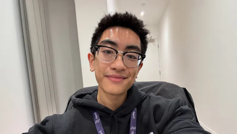 Duy-Anh wears a grey hoodie, black-rimmed glasses and a purple lanyard, standing in a corridor and smiling at the camera