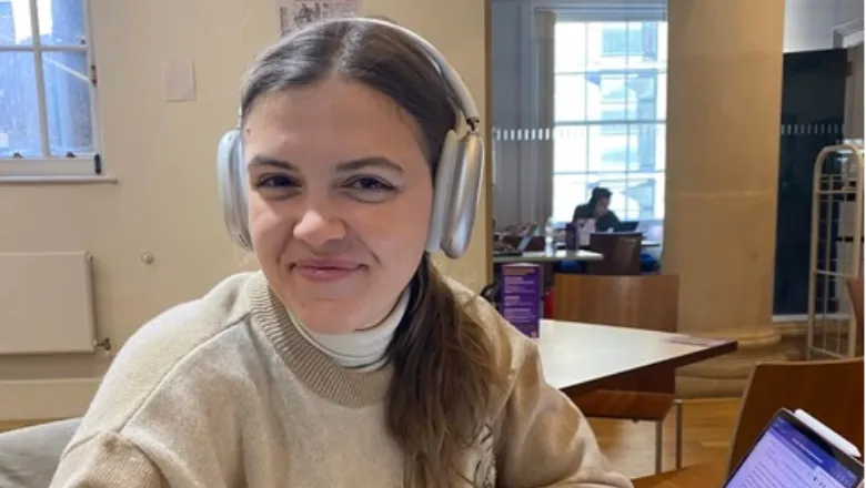 Giulia smiling at the camera, wearing headphones and using her laptop in Chapters Café on Strand Campus