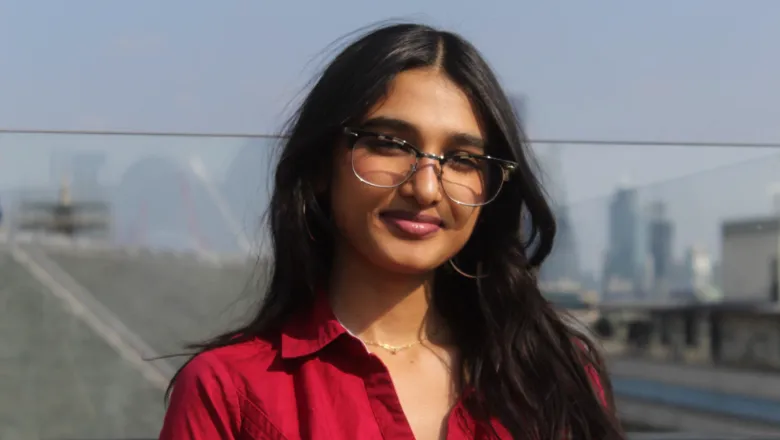 Neha wearing a red top, glasses and smiling at the camera with the sunny London skyline behind her