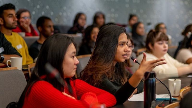 A person speaking into a microphone during a discussion in a lecture hall, while another person listens intently. The background shows several other attendees seated, some taking notes or listening, indicating the setting is a seminar or academic talk.