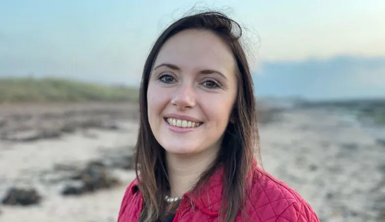 Professor Rebecca Goss faces the camera smiling, with a beach in the background