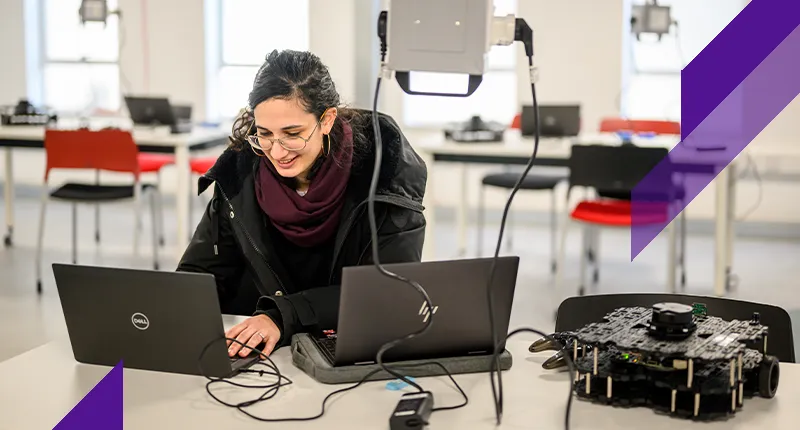 A young women works on two computers while a robotic device sits next to her.