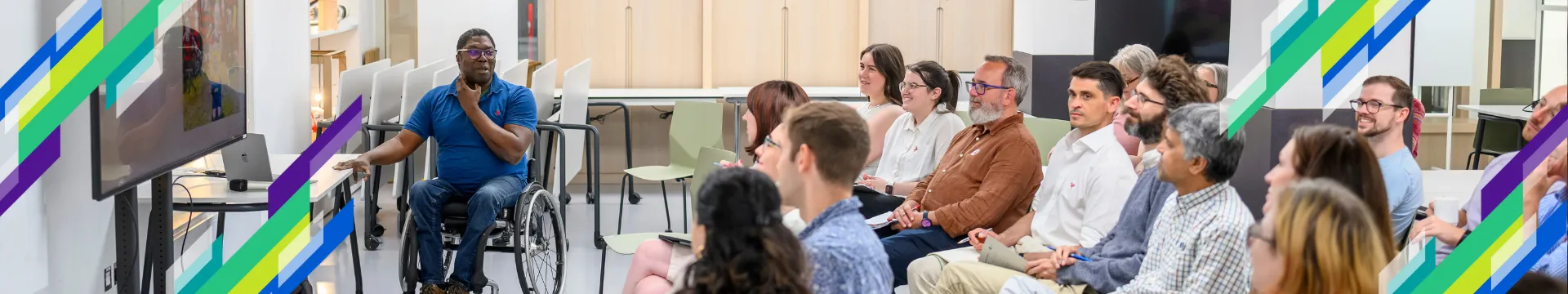 A lecturer in a wheelchair talks to an audience of seated adults in a brightly lit room