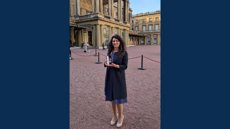 A woman in a black coat and blue dress holds her medal outside the palace.