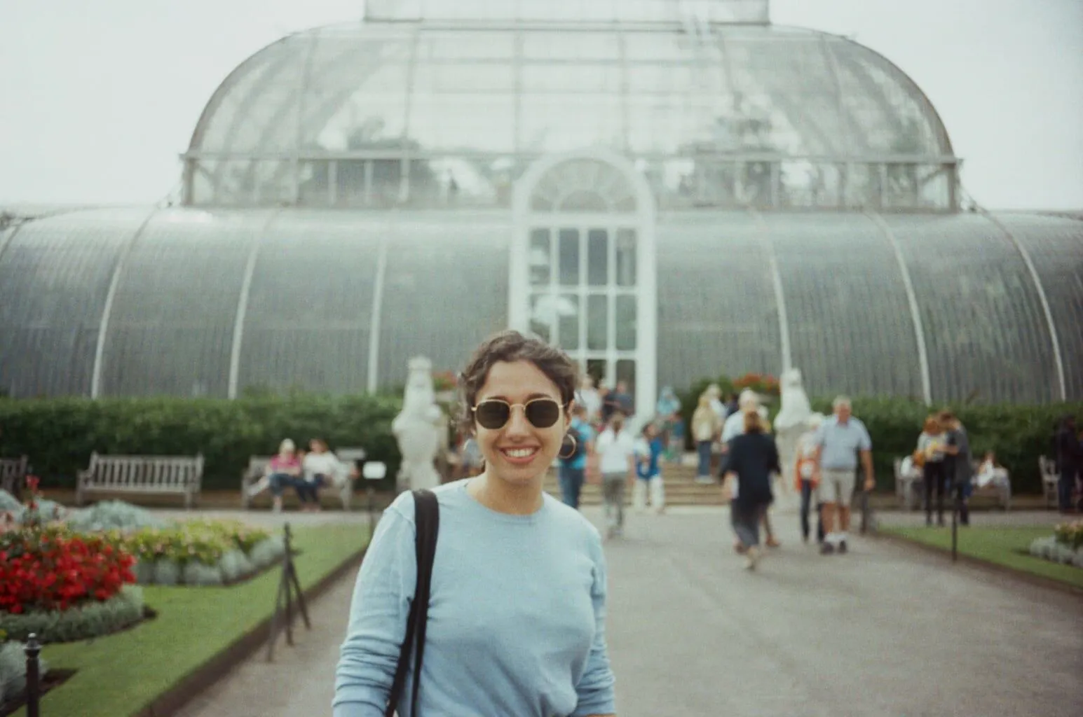 A person in a light blue jumper stands in front of the glass house at Kew Gardens. The film photo grain is visible.