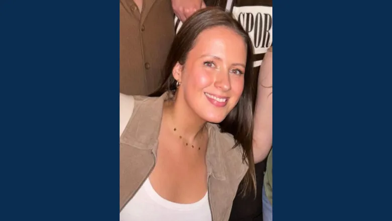 A young woman with long brown hair stands smiling wearing a tan suede jacket and white scoop top