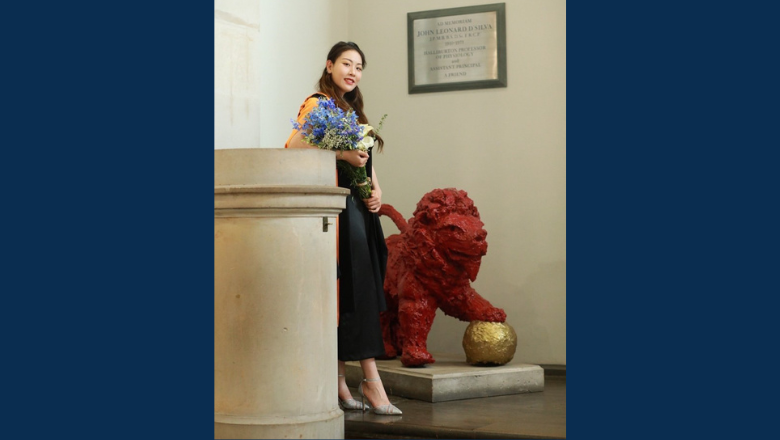 A woman stands smiling by a red lion statue which has its paw on a golden ball.