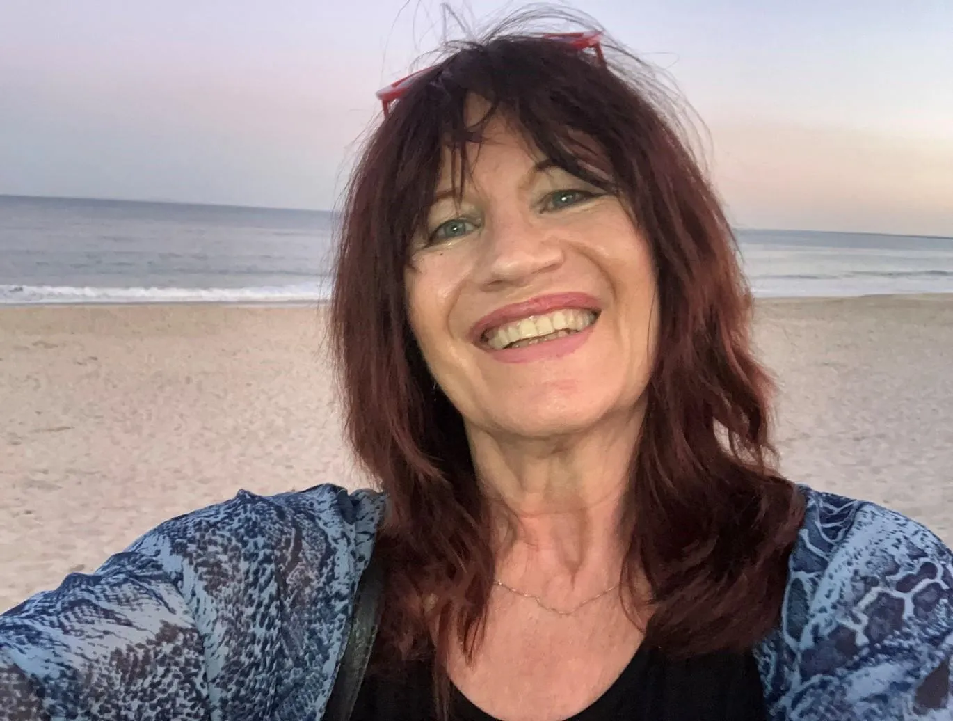 A woman with dark red hair smiles taking a selfie on a beach.