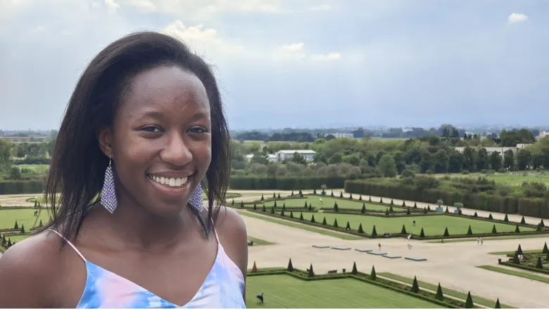 A woman smiles in front of a garden wearing a strappy blue and pink dress.