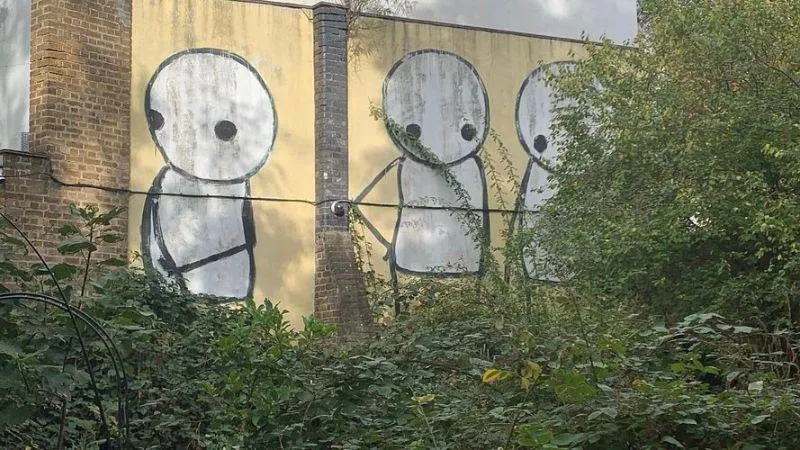 Street art on a brick wall depicting three stick figures with large eyes, partially obscured by surrounding greenery.