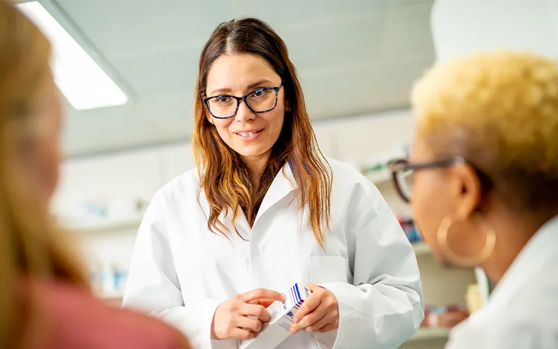 A person wearing glasses and a white lab coat holding a small box, in conversation with two other people