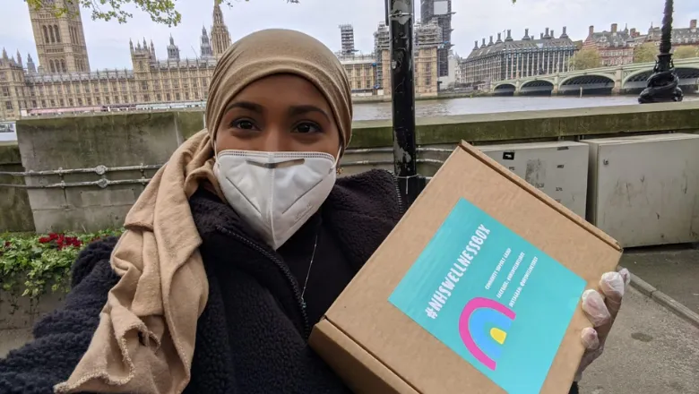 A person in a mask holds a care package for the NHS with the Houses of Parliament in the background