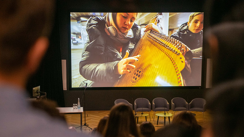 Audience watches Soldiers of Song. The image on a large screen shows a close-up of a woman playing a string instrument.