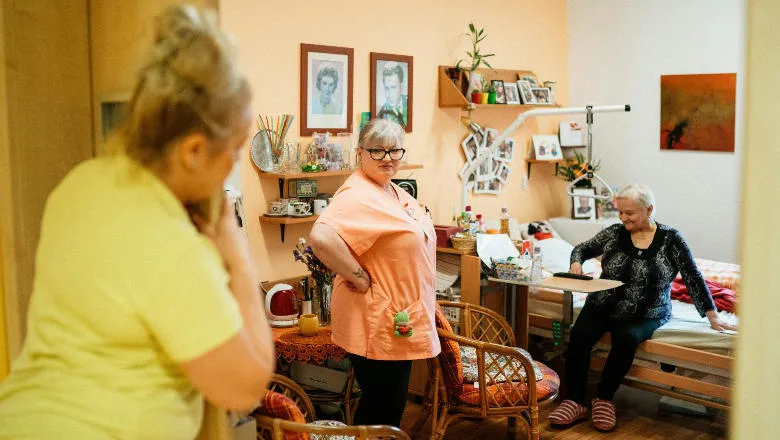 Person sitting on the edge of a bed with two carers nearby, one of them looking in from the door to the room