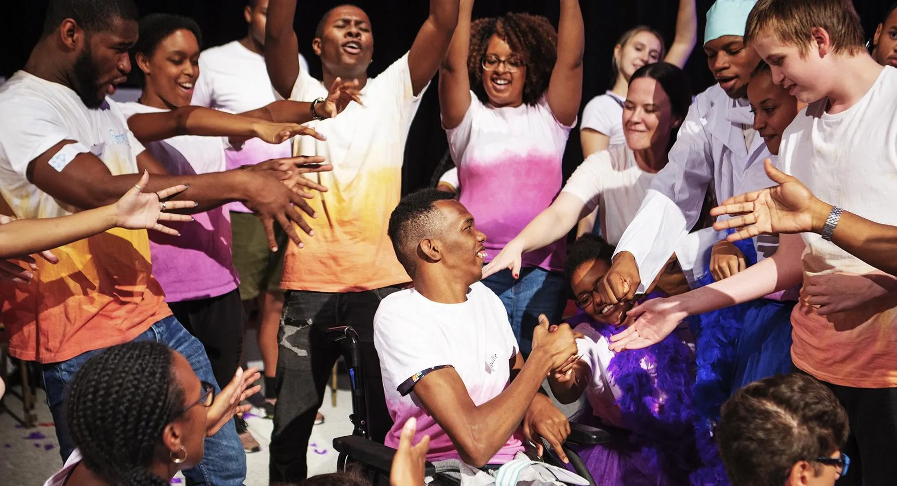 A person in a wheelchair surrounded by a group of happy people with their arms outstretched.