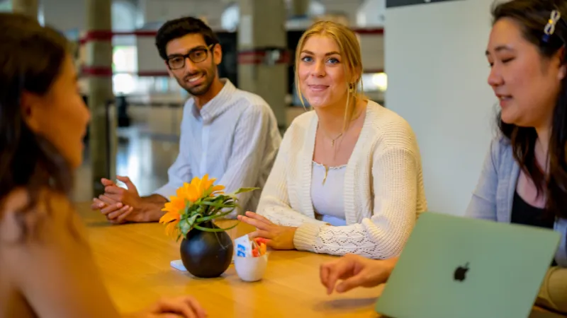 Four young adults sit around a table in conversation. A man with glasses smiles, while a blonde woman in a white sweater faces the camera. A laptop and a vase with a sunflower are on the table.