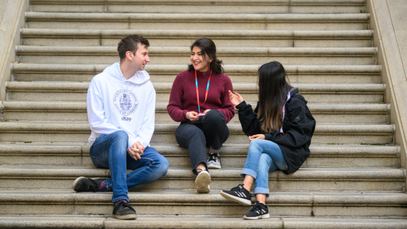 Three students sit on outdoor steps, casually chatting. One wears a white hoodie with a university logo, and the others wear jackets and sweaters.