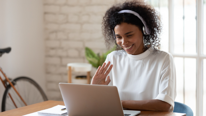 A Black female student wearing headphones, sitting at a table in front of an open laptop being on a call and smiling.