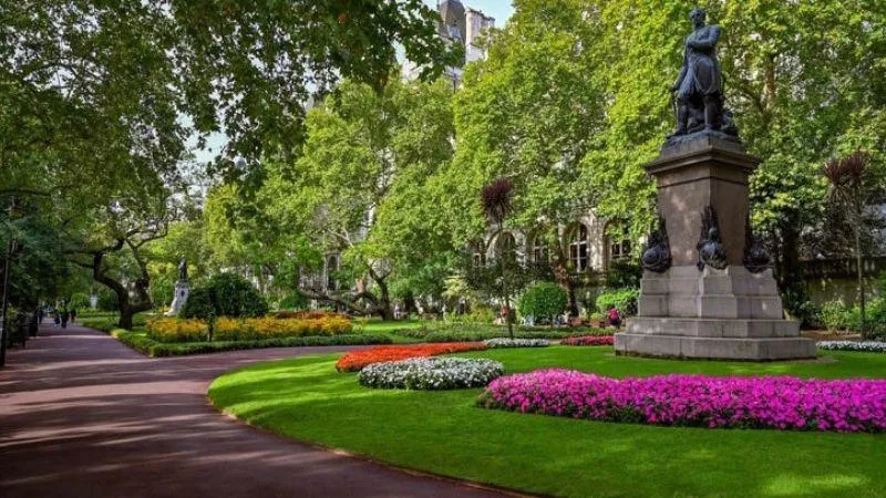 A lush park scene featuring a statue on a pedestal, surrounded by colorful flower beds and neatly trimmed grass, with tall trees lining a paved pathway on the left.