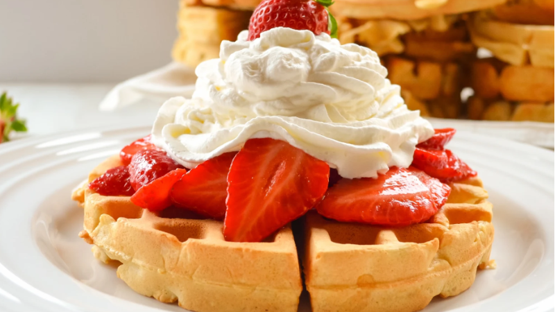 A close-up of a golden waffle topped with fresh sliced strawberries, a dollop of whipped cream, and a single whole strawberry on top. The dish is served on a white plate, with more waffles stacked in the blurred background.